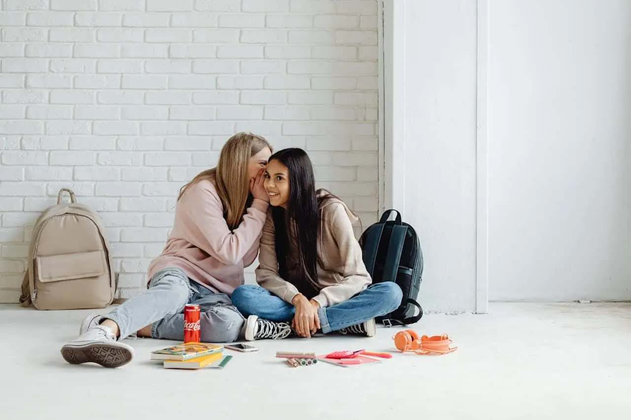 Two teen girls sitting on the floor and talking to each other with smiling face